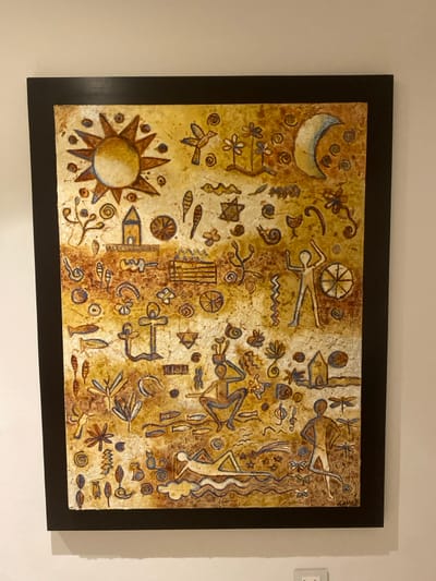 The artwork features a brown and gold textured background adorned with various symbolic motifs. Prominent symbols include a sun with rays, a crescent moon, stick figures in different poses, flowers, and geometric shapes. The figures and symbols are outlined with darker brown and hints of blue. The composition resembles ancient cave paintings or petroglyphs. The painting is encased in a thick, dark brown frame and mounted on a light-colored wall.