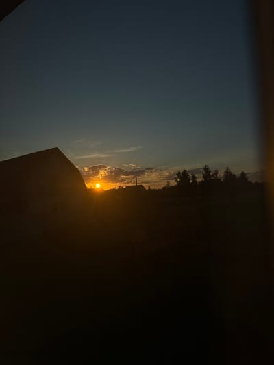 The image depicts a sunset scene in Chemerne, Ukraine. The sun is setting low on the horizon, casting a warm orange glow through scattered clouds. The sky transitions from a deep blue at the top of the frame to lighter hues of orange and yellow near the horizon. Silhouetted against the bright sky is a line of trees, their dark forms creating a stark contrast. In the foreground, the roofline of a building is visible, also in silhouette. The overall impression is one of tranquility and the end of a day. The lighting suggests it is either early morning or late evening. There is no visible text in the image.