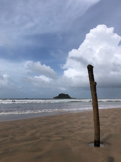 The image depicts a beach scene in Weligama, Sri Lanka. In the foreground, a weathered wooden post stands vertically in the wet sand, partially submerged in a small pool of water. The post is rough and textured, with visible knots and a string tied around its upper section. The sand exhibits rippling patterns from the receding tide.
In the midground, the ocean stretches out with moderate waves breaking near the shore. Several individuals are visible in the water, appearing to be swimming or surfing. A small, verdant island with a structure on top is situated in the distance, beyond the breaking waves.
The sky above is a dramatic mix of heavy, dark gray clouds and bright, fluffy white cumulus clouds. The lighting suggests it might be overcast or that a storm is approaching or has recently passed. The overall atmosphere is one of a coastal environment with signs of recent weather activity.