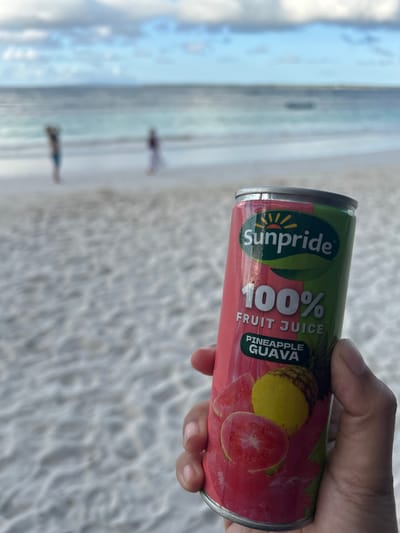 In the foreground, a hand holds a can of Sunpride 100% Fruit Juice, Pineapple Guava flavor, against a blurred background of a beach with gentle waves and a cloudy sky. Two figures are visible walking on the beach in the distance. The scene appears to be a tropical or coastal location.