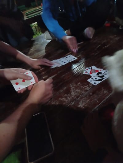 People are gathered around a wooden table playing cards. A hand holds the 3 of hearts. There are other scattered cards on the table, including face cards. A cell phone sits on the table. The location is potentially Gavrailovo, Bulgaria.