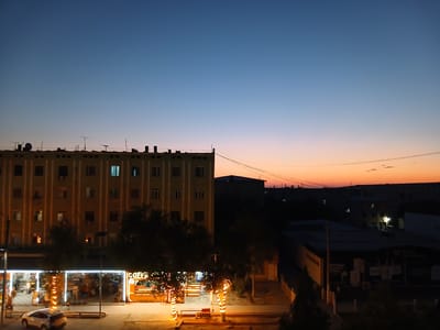 This image captures a twilight scene in Qorowul, Uzbekistan. A large, light-colored apartment building with numerous windows dominates the left side of the frame. Below the building, a street-level commercial area is illuminated, featuring a "COFFEE" sign and decorated trees wrapped in lights. A white car is parked on the street in front of the commercial establishments. In the background, other buildings and structures are visible, silhouetted against a gradient sky transitioning from deep blue at the top to hues of orange and pink near the horizon. Power lines crisscross the sky in the upper right portion of the image. The overall impression is an urban setting at dusk, with artificial lights beginning to illuminate the scene as natural light fades.