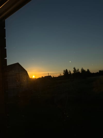 The image captures a sunset scene with a silhouette of a building in the foreground, against a gradient sky of dark blue transitioning to yellow and orange near the horizon. The sun is visible as a bright orb peeking over the horizon. Trees form a dark line against the lighter sky in the background. The lower portion of the image shows the edge of a structure, possibly a fence or deck, with a jagged top edge. There are specks of light, potentially dust or insects, scattered throughout the scene. The overall mood is serene and peaceful, typical of a sunset. The image is oriented vertically, with the horizon line running across the middle. No text is visible. The location context of Chemerne, Ukraine suggests a rural or semi-rural setting. The time of day is clearly sunset, indicated by the low position of the sun and the warm colors in the sky. The weather appears clear, allowing for a visible sunset.