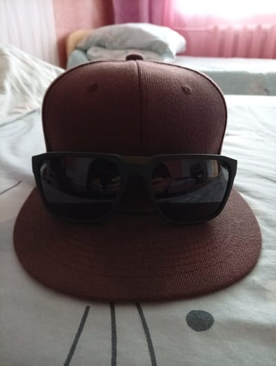 A close-up shot shows a brown baseball cap with sunglasses placed on top of its brim. The cap and sunglasses are positioned on a bed with a white and gray striped comforter. In the background, a pink curtain and a light blue blanket are visible, suggesting an indoor setting, likely a bedroom. There is no indication of Yanaul, Russia, in the image itself.
