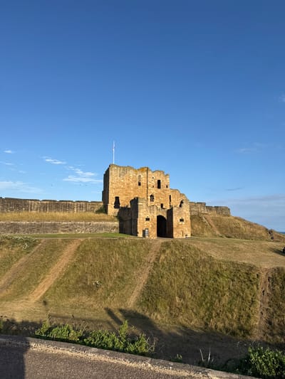 The image depicts a daytime outdoor scene featuring Tynemouth Castle and Priory, located in North Tyneside, United Kingdom.
The primary subject is a large, light brown stone castle structure situated on an elevated grassy embankment, occupying the mid-ground. The castle appears partially ruined, with visible battlements, multiple window openings, and a prominent arched entryway in its lower central section. A white flagpole, topped with a Union Flag, extends from the highest point of the main castle tower on the left. Stone defensive walls extend horizontally to the left from the castle, along the top of the embankment.
The embankment slopes downwards towards the foreground, covered in dry, patchy green and brown grass. Multiple narrow dirt paths are visible worn into the grassy slopes, indicating foot traffic. Long shadows are cast across the lower parts of the embankment and the immediate foreground, originating from the left side of the frame. The immediate foreground shows a grey, textured surface, possibly a paved path or low retaining wall, with sparse green vegetation growing along its edge.
The sky occupies approximately the upper two-thirds of the image, displaying a clear, bright blue hue with minimal cloud cover, predominantly on the right horizon. Several small, dark specks, consistent with birds, are visible in the sky to the left of the castle. A narrow strip of dark blue water, indicative of the sea, is visible along the far right horizon, beyond the castle embankment. A single indistinct, dark figure, possibly a person, stands on the upper grassy area to the far right, near the edge of the frame. A dark sign with white text is affixed to the wall near the castle's arched entrance.