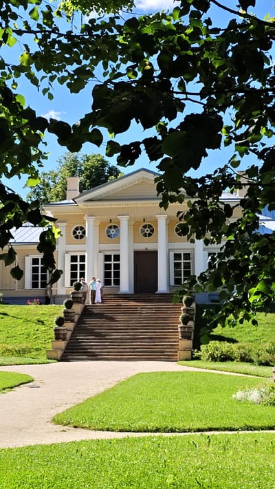 The image shows a neoclassical building with a central portico supported by four large white columns. The building is painted a pale yellow and features white trim and shutters on its windows. Above the windows are circular decorative elements. A wide, stone staircase leads up to the main entrance of the building, flanked by ornate planters with spherical topiaries. Two people, dressed in white, are standing on the steps, facing the building. The building is set in a lush green park with manicured lawns and trees. The sky is blue with a few scattered clouds. The scene appears to be set on a sunny day, likely in the late morning or early afternoon, given the strong sunlight and shadows. The overall atmosphere is peaceful and serene, suggesting a visit to a historical estate or museum in a park setting. The location is Vyborg, Russia, which is known for its historical architecture and parks. No visible text is present in the image.