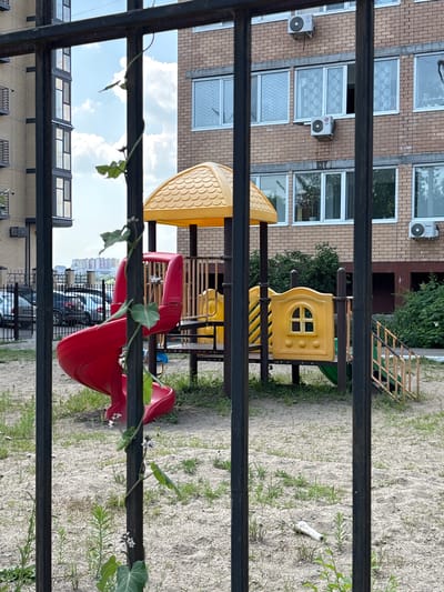 The image depicts a children's playground situated in the city of Blagoveshchensk, Russia, as observed through a black metal fence. The playground features a red spiral slide, a yellow play structure with a roof, and a yellow panel resembling a small house with a window. Green vegetation, including a climbing vine with small white flowers, is growing around the fence and the playground equipment. In the background, a multi-story brick building with air conditioning units and windows is visible, along with several parked cars behind the fence and a distant cityscape. The ground of the playground is covered in sand and sparse grass.
