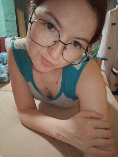 A young adult female is depicted in a close-up, chest-up shot, positioned as if reclining or lying down and propped on her forearms. She wears large, dark-rimmed polygonal glasses and has dark hair, with a neutral to slightly smiling facial expression. Her attire consists of a sleeveless top featuring a teal upper section and a light grey lower section, complemented by a thin necklace with a small pendant. Her right forearm is crossed over her left. The immediate surface she rests upon is light brown. In the background to the left, crumpled blue and white patterned fabric is visible, along with a greenish-blue textured surface and a dark, vertical rectangular object. To the right, a white garment with blue accents, possibly a uniform, is hanging or draped. The location is Старый Шагирт, Russia.