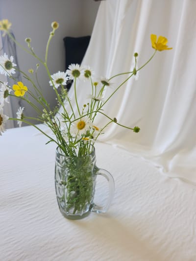 A bouquet of daisies and yellow buttercups are arranged in a glass mug. The mug has a handle and a textured pattern. The bouquet is placed on a white, textured surface, possibly a tablecloth. In the background, there is a white, draped fabric, which appears to be a backdrop, and a dark object, potentially furniture, out of focus. The scene is brightly lit with soft shadows cast by the bouquet. There is no indication of a specific location in Gatineau, Canada.