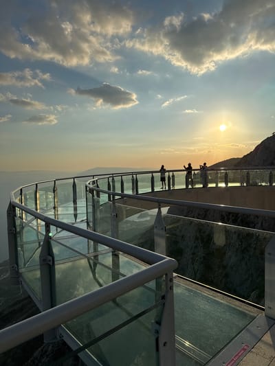 The image depicts a glass-bottomed observation walkway on a mountain at sunset. Several people are visible on the walkway, silhouetted against the bright sun. The walkway is constructed with glass panels and metal railings, offering a panoramic view of the landscape below and the distant horizon. The sky is filled with dramatic clouds, illuminated by the setting sun. The location is identified as the Biokovo Skywalk in Općina Tučepi, Croatia, a popular tourist attraction offering stunning views of the Adriatic coast and islands.