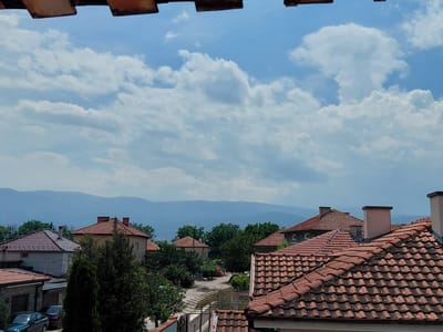 This image, taken in Krumovo, Bulgaria, presents an elevated daytime view over a village landscape. The upper portion of the frame features a clear blue sky interspersed with scattered white cumulus clouds, varying in density and form. A blurred, dark red-brown structure, consistent with an overhead roof or eaves, partially frames the top edge of the image.
The mid-ground and foreground are dominated by numerous residential buildings, characterized by terracotta-red, interlocking tile roofs. These roofs display various configurations, including gabled and multi-pitched designs, with visible wear and discoloration on some tiles. Several chimneys, primarily light beige or pinkish-brown in color and rectangular in shape, project from the rooftops.
Lush green trees and dense foliage are abundant throughout the scene, partially obscuring the houses and creating a verdant canopy. Coniferous and deciduous tree types are present. In the distant background, a range of mountains or hills appears hazy and blue, indicative of atmospheric perspective.
An unpaved, light-colored road or path winds through the houses in the mid-distance. In the lower-left section, two automobiles are partially visible: a dark-colored sedan parked in front of a garage and a second dark vehicle positioned behind it. A metal grid-like structure, possibly a clothesline or trellis, is visible in the lower-mid section, spanning between houses and trees. No human figures are present within the frame. The overall light suggests a sunny day with intermittent cloud cover.