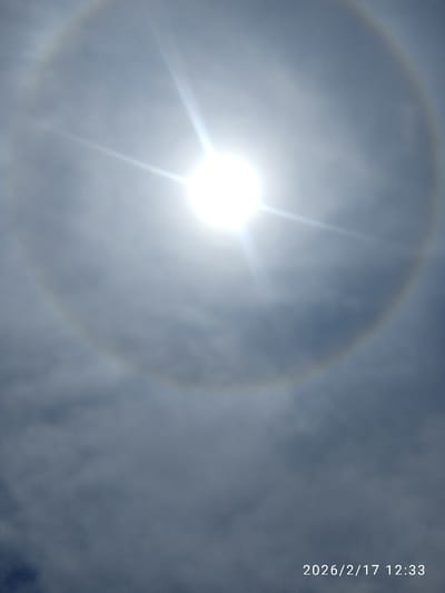 The image displays a solar halo encompassing a bright, central sun with pronounced sunburst rays, set against a partially overcast, grayish-blue sky. The halo appears as a wide, circular arc of light. No human figures, structures, or man-made objects are visible within the frame, indicating an unobstructed view of the sky. The scene is situated in Athi River ward, Kenya. A digital timestamp "2026/2/17 12:33" is visible in the lower right corner.