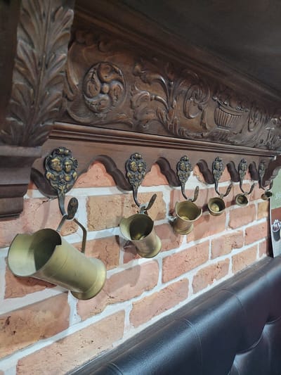 A close-up, eye-level shot captures a wooden shelf with ornate carvings above a brick wall. Several brass hooks with decorative facial motifs hang from the shelf, each supporting a small brass vessel. The vessel on the far left is a cylindrical cup tilted downwards. The other vessels appear to be smaller bowls or cups, some containing what looks like small, round objects, possibly balls or coins. The brick wall has a textured, rustic appearance with white mortar. Below the wall, the edge of a black leather bench is visible. The overall scene suggests an interior space, possibly a bar or restaurant, with a decorative and vintage aesthetic.