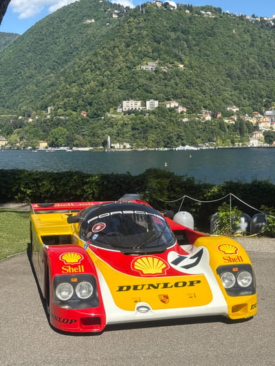 The image showcases a Porsche 962C race car, liveried in the Shell-Dunlop racing colors. It is parked on a paved area, presumably near Lake Como, Italy. The background features Lake Como, surrounded by a densely wooded hillside with scattered buildings. The car's exterior is predominantly yellow, red, and white, with prominent Shell logos and Dunlop lettering. The cockpit is covered and reflects the surrounding scenery. The scene is well-lit, indicating a sunny day. The car appears to be stationary.
