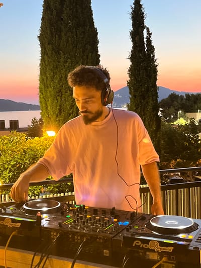 A DJ wearing headphones and a pink t-shirt is playing music on a Pioneer DJ setup at dusk in Pržno, Montenegro. The DJ is standing in front of the equipment, with their hand on a turntable. The background shows a scenic view of the Adriatic Sea and surrounding hills, with cypress trees framing the scene. The sky is a gradient of orange and blue, indicating the sunset. The DJ booth is set up on a balcony or terrace, with a railing and plants visible. The overall atmosphere is relaxed and summery.
