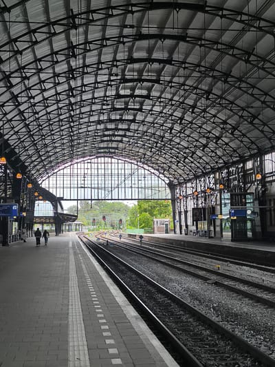 The image depicts the interior of a train station in Haarlem, Netherlands, characterized by its grand, arched metal roof structure. The roof is supported by a complex network of beams and crossbars, with corrugated metal sheeting forming the canopy. Numerous orange-tinted lamps are suspended from the roof. A large, arched glass window dominates the far end of the station, offering a view of greenery and trees outside.
In the foreground, a tiled platform runs alongside multiple sets of train tracks. The platform is marked with tactile paving and white lines. Two individuals are walking away from the camera on the left-hand platform, appearing to be heading towards the far end of the station. On the right-hand side, the train tracks recede into the distance. Signal lights are visible above the tracks, indicating their operational status. Further down the platform, information displays and glass-enclosed structures are present. The overall atmosphere is that of a functional and architecturally notable transportation hub.