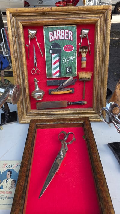 The image depicts a stall at an outdoor market, likely a flea market or antique fair, with items displayed for sale. The primary focus is a framed collection of vintage barber tools. The collection is mounted on a red felt background within a golden, ornate frame. Items visible include a manual hair clipper, scissors, a small glass bottle, a straight razor with a wooden handle, a sharpening strop, and two shaving brushes. A vintage "Barber" sign, featuring a barber pole design and the text "EST 1934 Enjoy!", is also prominently displayed.
Below this framed display, a separate, smaller wooden frame with a red felt interior holds a pair of large, antique scissors. To the left of the framed items, partially visible, is a vintage silver hair dryer. Further to the left and bottom, a booklet or small book with illustrations of people and text in Spanish, possibly related to rural life or a magazine, is visible. The background suggests an outdoor setting with a vehicle and trees. The overall impression is one of vintage Americana or historical grooming tools.
While the image itself does not provide explicit geographical markers for Almuñécar, Spain, such markets are common in tourist towns and historical centers. If this stall is indeed in Almuñécar, it would likely be situated in or near the town's historic center, such as the Plaza del Ayuntamiento or along the main streets leading to the beach or the Castillo de San Miguel. The presence of vintage items and crafts is typical of such locations.