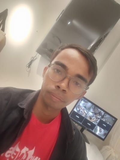 This media file captures a man in a close-up selfie, likely taken indoors. He is wearing glasses with clear, round frames and a dark collared shirt over a red t-shirt. The red t-shirt features a white graphic design. The background is dominated by a white ceiling and a large, dark rectangular object, possibly a monitor or screen, mounted on the wall. This screen displays multiple camera feeds, suggesting a surveillance system is in place, possibly in a retail or office environment. There are also wires visible, connected to the screen, and a bright light source is prominent in the upper left quadrant of the image. The man's expression is neutral. The image quality is somewhat grainy and appears to be taken with a phone camera. The location context provided is Larangan, Indonesia.
