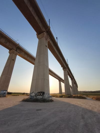 This low-angle photograph captures two parallel concrete railway viaducts in the Mate Yehuda Regional Council, Israel, during either sunrise or sunset. The sky transitions from a deep, clear blue at the top to a warm, golden orange near the horizon on the left.
The foreground is dominated by the tall, tapering concrete piers of the bridges. The most prominent bridge, positioned towards the right, features several large, light-grey concrete piers supporting a wide, flat deck equipped with overhead electrical lines and dark railings. A second, similar concrete bridge is visible further left and at a slightly lower elevation, also featuring multiple piers and a deck with blue railings and overhead lines.
Beneath the bridges, the ground is a dry, light-brown dirt surface, traversed by a path or road. Long, distinct shadows from the piers stretch across the ground, indicating a low sun angle. Sparse, dry vegetation, including grasses and small shrubs, grows around the bases of the piers and along the dirt path.
Graffiti is visible on the lower sections of the concrete piers. On the closest pier to the right, a black rectangular tag containing white "XXX" and other symbols is present, with the text "FUCK IRAQ" written below it. On a pier to the far left, a blue and white design, possibly resembling an Israeli flag, is visible alongside Hebrew text. A white vehicle is partially visible behind the rightmost pier, parked on the dirt.
The background shows a rural landscape with agricultural fields in various shades of green and brown, stretching towards a hazy distant horizon where faint structures can be discerned. The overall illumination is soft and warm, characteristic of the golden hour.