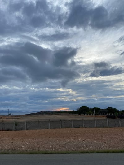 The image depicts a cloudy sky over a barren landscape. In the foreground, there is a gray asphalt road. Behind the road, a rough, dirt ground is visible, with sparse, dry vegetation. A low wall made of concrete blocks separates this area from a more distant, dry, grassy field. In the background, there are some trees and hills. The sky is filled with dark, gray clouds, with patches of lighter sky peeking through, suggesting either sunrise or sunset. There are no people or animals visible in the image. The scene appears to be an undeveloped area, possibly on the outskirts of a city.