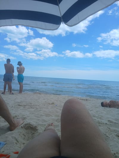 A first-person perspective shot captures a sandy beach with the sea and sky in the background. In the foreground, a person's legs are visible, partially covered in sand. To the right, a young child lies on the sand, appearing to be asleep. Further back, two young men are standing near the water's edge, facing away from the camera. One man wears patterned swim trunks, and the other wears blue swim trunks and a hat. A striped umbrella shades a portion of the scene. The location is identified as Osenovo, Bulgaria.
