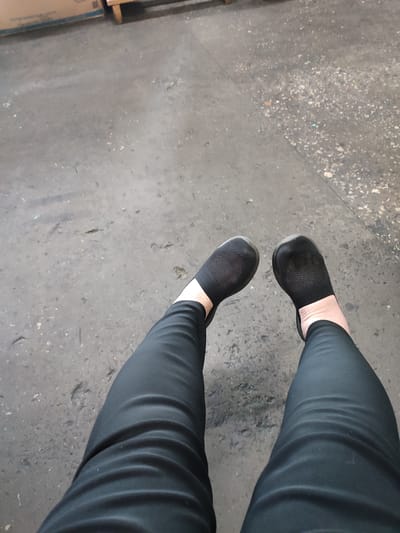 The image displays a pair of legs from the knees down, clad in black leggings and black slip-on shoes. The legs are positioned on a rough, concrete-like surface, appearing to be outdoors. The background is a textured gray ground with scattered debris. To the upper left of the image, a portion of a cardboard box is visible. The overall scene suggests an urban or industrial setting. There are no people or animals clearly identifiable beyond the subject whose legs are depicted. The lighting is even, indicating it is daytime. The image appears to have been taken from a slightly elevated perspective, looking down at the subject's legs. There are no specific landmarks or elements that definitively place the location in Narowlya, Belarus, as the environment is generic.
