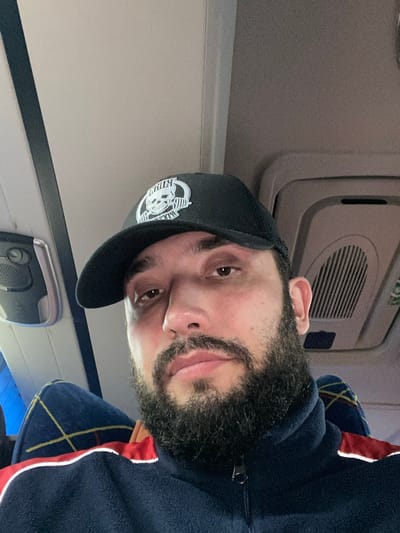 A man wearing a black baseball cap with a white logo, a dark blue fleece jacket with red and white trim, and a beard is sitting in a vehicle. The cap has a graphic of a skull and barbells. The man is looking towards the camera. The background shows the interior of the vehicle, including seats and overhead compartments. The man is sitting on the left side of the frame, with his head and shoulders visible. The setting appears to be indoors, likely in a bus or train, given the overhead fixtures and the design of the seats. There are no other people visible in the image.
