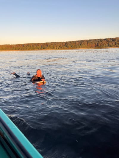In Gromovo, Russia, a person in a wetsuit floats in dark water, holding onto a small board. The person is facing the camera, with their body submerged from the chest down. Their legs are bent and positioned behind them, with feet visible above the water. The surrounding water is calm, with ripples and light reflections. In the background, a dense forest lines the horizon under a clear sky with a warm glow near the treeline, suggesting it is either sunrise or sunset. A portion of a teal-colored boat is visible in the lower left corner of the frame.