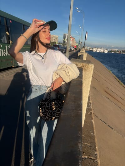 A young woman with long brown hair, wearing a white t-shirt, light blue jeans, a baseball cap, and a leopard-print handbag, stands on a waterfront promenade in Yoksólovo, Russia, on a sunny day. She holds her hand up to her forehead, shielding her eyes from the sun. To her left is a green bus, and to her right is a body of water with several white boats docked. In the background, city buildings and bridges are visible. The promenade is made of concrete, with a low wall separating it from the water.
