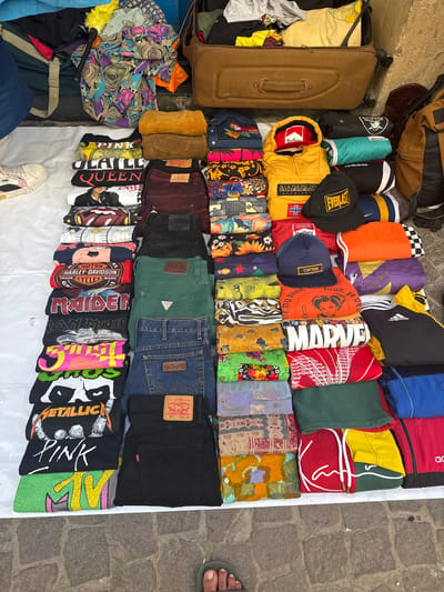 A street market stall in Essaouira, Morocco, displays a variety of folded clothing items, primarily t-shirts and jeans, laid out on a white cloth on a cobbled street. The stall is set up outdoors, with a suitcase and bags visible in the background, suggesting a commercial activity. The clothing includes graphic t-shirts with band logos like "Metallica" and "Maiden," as well as brand names such as "Guess" and "Levi's." There are also items with colorful patterns and designs. One person's feet, wearing a sandal, are visible in the lower right corner of the frame, indicating their presence at the stall. The lighting suggests daylight.