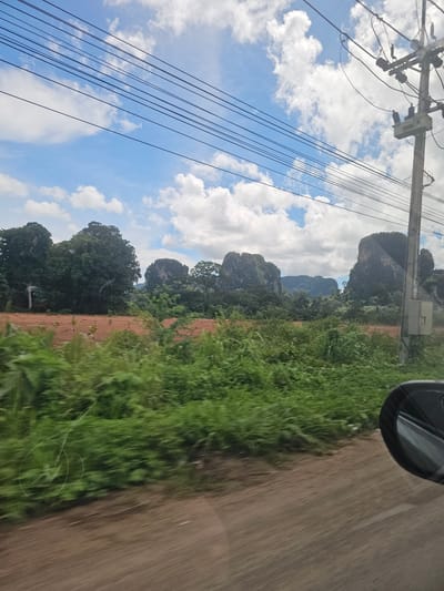 The image captures a scenic view of Ban Thung, Thailand, featuring a blend of natural and man-made elements. In the foreground, a dirt road runs along the right side, with lush green vegetation bordering it. The vegetation is in motion blur, indicating movement, likely from a vehicle. A side-view mirror of a car is visible on the far right of the frame.

Further into the scene, a reddish-brown field is dotted with small, young trees, suggesting recent planting or cultivation. Beyond this field, a dense forest stretches across the landscape, with several imposing, rocky karst formations rising majestically from the trees. These formations are a prominent geological feature of the region.

Overhead, a bright blue sky is adorned with scattered white cumulus clouds. A complex network of power lines and a utility pole are visible in the upper right quadrant of the image, indicating the presence of infrastructure. The overall scene is one of rural beauty, characterized by the dramatic landscape and the interplay of natural elements with signs of human presence.