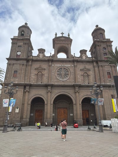 The image shows the Las Palmas Cathedral, also known as the Cathedral of Santa Ana, located in the Vegueta neighborhood of Las Palmas de Gran Canaria, Spain. The grand facade of the cathedral, constructed from dark stone, dominates the frame. It features two prominent bell towers flanking a central section with a large rose window. Multiple arched doorways and windows are visible.
In the foreground, a paved plaza stretches out. Several people are present. In the center of the plaza, a person wearing a pink shirt and green shorts is facing away from the camera, looking towards the cathedral. To the left, a street lamp with multiple globes stands tall. Banners with images and text are attached to the lamp post and the cathedral's exterior.
Further into the plaza, near the entrance of the cathedral, a small red forklift is parked. A white van is visible to the right, behind a lamppost, and a stack of white chairs is also present in that area. Several other individuals are walking around the plaza, some near the entrance and others in the distance. The sky above is partly cloudy.