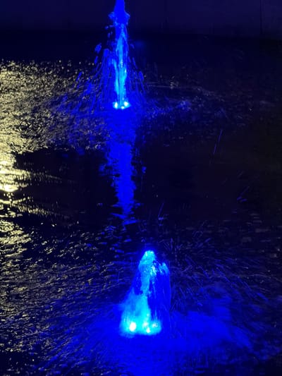 The image displays a vibrant and dynamic scene dominated by intense blue light against a dark background, likely captured at night or in an artificially darkened indoor setting. Two prominent sources of bright blue light are visible: one on the far left, appearing as a focused jet or beam, and another on the right, presenting as a broader, more dispersed glow.
Surrounding these primary light sources, numerous streaks, specks, and splashes of blue light suggest the energetic movement of liquid or gas, possibly water, mist, or some other fluid. This creates a texture of illuminated particles across the frame. Towards the bottom and extending slightly to the right, a surface, most likely water, reflects and diffuses a yellowish-white light, providing a strong contrast to the predominant blue and black palette.
The activity depicted appears to be a light and water display, possibly an illuminated fountain, a specialized car wash facility, or a modern artistic installation where light is used to highlight fluid motion. No people are visible, and there is no discernible text in the image. The scene is characterized by its dramatic lighting and the sense of movement conveyed by the illuminated spray. The location context provided is Osijek, Croatia.
