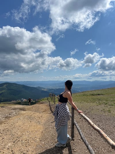 In the city of Chorna Tysa, Ukraine, a young woman stands on a dirt path, leaning on a rustic wooden fence. She wears a black crop top, light blue pants, a plaid shirt tied around her waist, and a black baseball cap. Her hair is in a braid that hangs over her shoulder. She looks out at a breathtaking landscape of rolling green hills and distant mountains under a bright blue sky dotted with fluffy white clouds. Further down the path, another person is visible, appearing to be running. The environment suggests a mountainous or hilly terrain, possibly a hiking trail or a scenic overlook. The ground is covered in dirt and small stones, with patches of dry grass on the slopes. The wooden fence is weathered and appears to be made of rough-hewn logs and branches. The scene is captured in daylight, with clear visibility.
