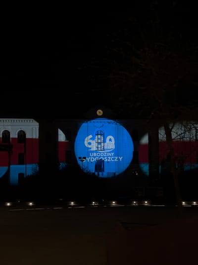 A building in Bydgoszcz, Poland, is illuminated at night with a blue light projecting the words "Urodziny Bydgoszczy" and the number "69" onto its facade. The building has a clock on its side. Above the blue light, the building has white, red, and blue sections that appear to be projected onto it as well. A tree is visible in the upper left corner. A row of lights runs down the side of the building on the right.