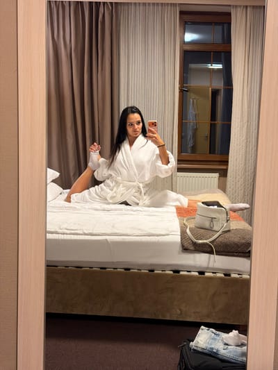 A young woman with long dark hair is performing a split on a hotel bed, wearing a white bathrobe and white socks. She is taking a selfie with her smartphone. A white handbag is placed on a folded blanket next to her. A suitcase with folded jeans and a white cloth is on the floor in the foreground, visible in the reflection of a mirror. The room has curtains, a window with a view of another room, and a radiator. The location is a hotel room in Svetlogorsk, Russia.