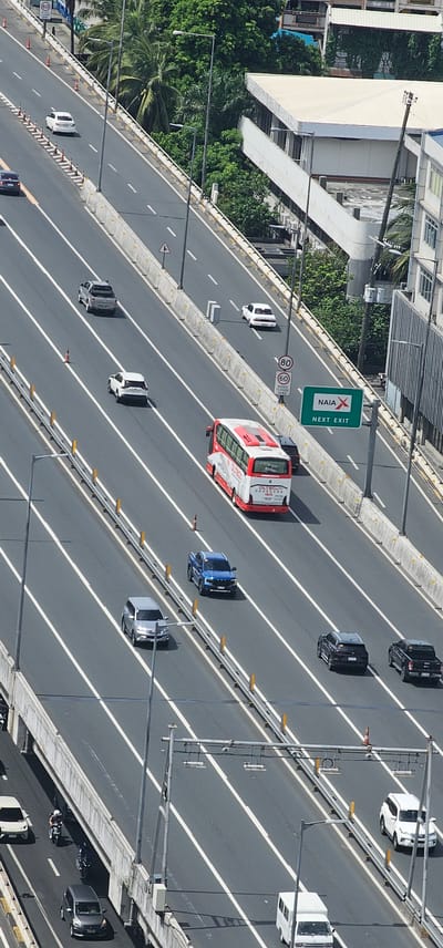 This aerial photograph captures a highway in Makati, Philippines, under daylight. The multi-lane highway features a mix of vehicles, including cars, a bus, a pickup truck, and motorcycles. White lane markings divide the asphalt road, with concrete barriers and metal railings separating the lanes and the roadside. Lush green trees and buildings are visible on the right side of the highway. A speed limit sign of 80 and 60 is posted. A green directional sign indicates "NAIA X NEXT EXIT." The perspective is from above, looking down at the traffic flow.