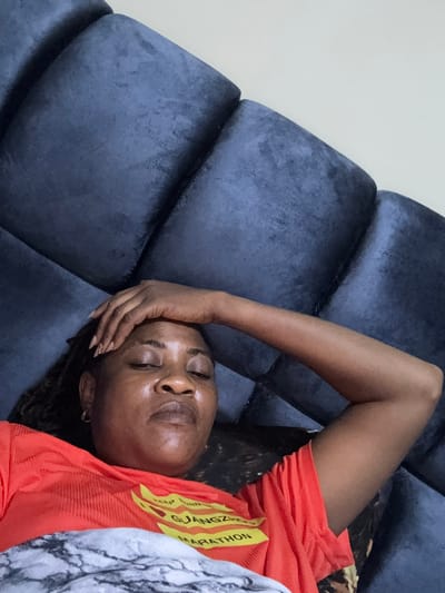 In a bedroom in Ajara, Nigeria, a woman rests on a large, dark blue tufted headboard, her eyes closed and her hand on her forehead. She is wearing an orange t-shirt with yellow lettering that reads "KEEP RUNNING GUANGZHOU MARATHON". Her arm is bent at the elbow and resting on the headboard. The lighting suggests it is either early morning or late evening, as the room is dimly lit. The woman appears to be in a state of rest or repose.