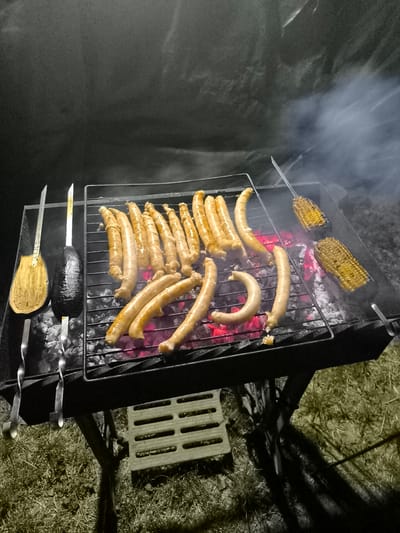 In Țipletești, Moldova, a barbecue is underway. The scene is illuminated by the red glow of hot coals and the dim light of dusk, with smoke rising from the grill. A metal grill rack sits atop a charcoal brazier, filled with glowing embers. On the grill, numerous sausages are cooking, some arranged in rows and others curved. To the left of the sausages, two pieces of eggplant are skewered and positioned on the coals. To the right, two ears of corn on skewers are also being grilled, their kernels appearing yellow and slightly charred. The surrounding ground is covered in dry grass, and a shadow of the barbecue structure is cast upon it. The overall atmosphere suggests a nighttime outdoor meal preparation.