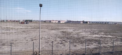 The image depicts an outdoor scene under a clear, pale blue sky, suggesting daytime with fair weather. The foreground features a chain-link fence, with metal posts and a coiled cable visible near the bottom right. Beyond the fence lies a large, sparsely vegetated, arid expanse of dry earth and scrubby plants.

In the middle ground, a tall, slender lamppost stands prominently on the left side, topped with a rectangular light fixture. Further in the background, a row of shipping containers, some colored red, green, and grey, are neatly arranged. Beyond the containers, a long, low-rise building complex with red roofs stretches across the horizon. The architecture of these buildings suggests a residential or communal living facility. In the far distance, more industrial or infrastructural elements, including what appear to be power lines or communication towers, are visible.

The overall setting appears to be an undeveloped or industrial area on the outskirts of a settlement, possibly a port or logistics hub, given the presence of shipping containers. The arid landscape and sparse vegetation are indicative of a dry climate. No people are visible in the image, and there is no discernible text. The scene conveys a sense of an industrial or administrative zone within a somewhat desolate landscape.