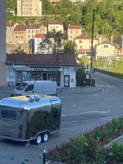 The image captures a street scene in the city of Saint-Claude, France. In the foreground, a polished, retro-style silver trailer with two white wheels and a visible license plate reading "WW-299-DS" is parked on asphalt. Two doves are visible on the ground near the trailer. Behind the trailer, a white delivery van is parked in front of a small commercial building with a sign that reads "TABAC PRESSE." The building has a reddish-brown, tiled roof. Further in the background, a gently sloping road curves to the right, bordered by a black metal railing and lush green trees. Several residential buildings with varying architectural styles and muted colors are situated on a hillside overlooking the street. The architecture suggests a European urban setting, with some buildings featuring balconies and multiple windows. The overall lighting suggests it is daytime, likely late afternoon due to the warm tone of the light on the buildings.