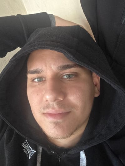 The image displays a close-up, vertical portrait of a male individual. He has light-colored eyes and short facial hair, including a mustache and stubble on his chin and jawline. The individual is wearing a black hooded sweatshirt with the hood pulled over his head, framing his face. A partial white logo, identified as "Lonsdale London," is visible on the lower left chest area of the sweatshirt. A black zipper and a white drawstring are present on the front of the garment. The individual is looking directly forward, towards the camera, with a neutral expression. The background is out of focus, showing light-toned, unidentifiable surfaces, suggesting an indoor environment. The specific location within the city of Brusen, Bulgaria, cannot be determined from the visual information contained in the image.