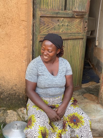 A Black woman wearing a gray t-shirt and a colorful wrap skirt sits on a step in front of a weathered green metal door. The door has vertical slats and a decorative pattern of diamond shapes. The woman has a black headscarf on, and she is smiling and looking to her right. Her hands are clasped in her lap. To her left, a large silver pot with a lid is visible. The wall next to the door is made of rough, light brown stucco, and to the right of the door, a narrow opening reveals a dark interior with a blue rug. The scene appears to be an outdoor setting, possibly in a residential area in Riyom, Nigeria.