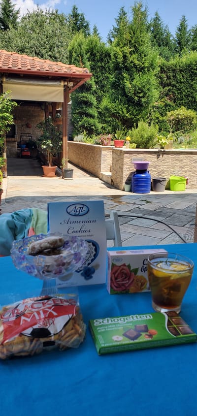 The image captures a sunny outdoor scene, likely in Νέα Σάντα, Greece, with a bright blue tablecloth laid out on a tiled patio. The table is laden with a small spread of snacks and drinks. A glass of amber liquid, possibly tea with lemon, sits next to a box of "Agi Armenian Cookies" adorned with roses. Further along, a green box of "Schogetten Alpine Milk Chocolate with Hazelnuts" is visible, alongside a bag of "Crackers Mix Salty". The background features lush green trees and a section of a terracotta-tiled roof, suggesting a private residence or garden area. The overall impression is one of a casual, pleasant outdoor gathering on a bright day. There are no people visible in the frame, but the presence of snacks implies an activity or event. The lighting indicates it is daytime.