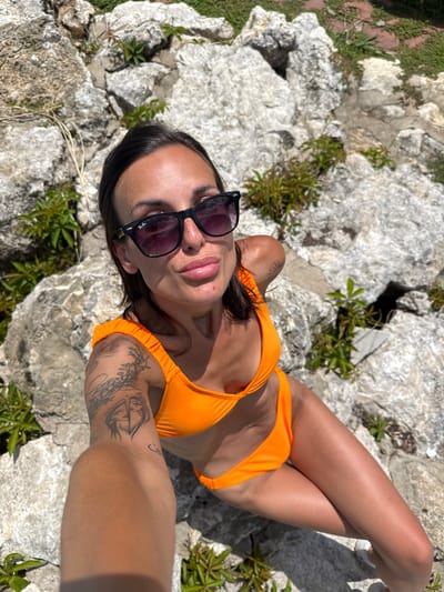 A young woman with dark hair, wearing sunglasses and an orange bikini, is sitting on rocks in a natural outdoor setting. She has tattoos on her arm and is looking at the camera with her lips slightly parted. The background consists of large gray rocks and some green vegetation, suggesting a rocky, natural landscape. The image is taken from a high angle, looking down at the woman. The location is not specified within the image itself, and the mention of "Grivitsa, Bulgaria" is external information.