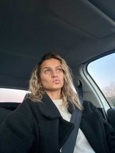 A woman with wavy blonde hair and green eyes sits in a car wearing a black coat and a white sweater. A seatbelt crosses her chest. She is looking upwards with slightly pursed lips. The interior of the car is visible, including the ceiling and a window showing trees and sky outside.