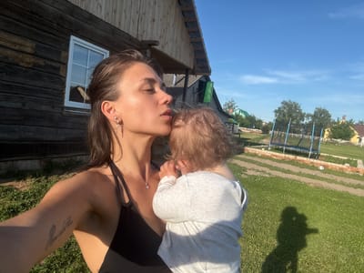 A woman is holding a baby and kissing its head in front of a wooden house. The woman is wearing a black tank top and a silver necklace. The baby is wearing a light gray shirt. The house is made of dark wood and has a white-framed window. The background shows a grassy yard, a trampoline, and other houses. The sky is clear and blue. The image appears to be taken in a rural area. The location is Зажэвіцкі сельскі Савет, Belarus.