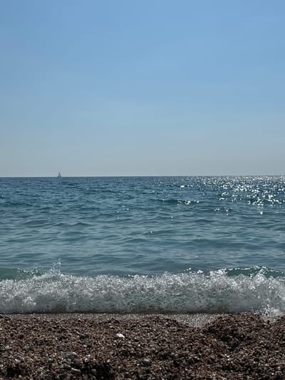 This media file depicts a serene seascape with a focus on the meeting of water and shore. The scene is divided into three distinct vertical sections. On the left, a clear, pale blue sky occupies about half of the frame, transitioning to a slightly lighter hue towards the top. In the middle, a body of calm, rippling water stretches across the frame, catching the sunlight and shimmering with bright highlights. A lone sailboat with a triangular sail is visible in the distance, a small white silhouette against the blue expanse of the sea. The rightmost section of the image is a close-up view of a pebble beach. The pebbles are dark brown and grey, appearing wet and glossy, with many reflecting the sunlight. A gentle wave is breaking onto the shore, creating a frothy white line of foam that partially covers the pebbles. The overall impression is one of tranquility and natural beauty. The lighting suggests it is daytime, possibly mid-morning or afternoon, under fair weather conditions. The location cues, such as the pebble beach and the calm sea, are consistent with a coastal environment. There is no visible text in the image.