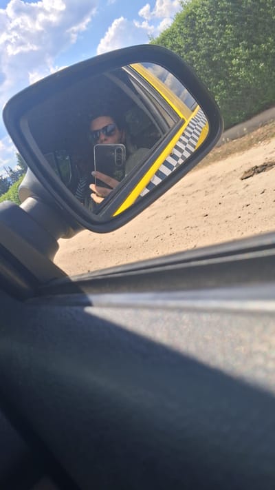 A person is taking a selfie in the side mirror of a yellow taxi. The taxi has a checkered pattern on its side. The person is wearing sunglasses and holding a smartphone. The background shows a dirt road and greenery. The sky is blue with some clouds.