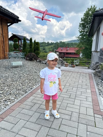 A young child stands on a grey paved walkway, angled slightly to their right (our left). The child wears a white baseball cap with a subtle floral design, a white short-sleeved t-shirt with pink text that reads "Sip Bella" and a yellow lemon graphic, bright pink shorts, and white sandals featuring small yellow cartoon characters. Their expression is neutral to slightly curious.
To the child's left, the walkway borders a gravel path. In the background, beyond the gravel, are various green trees and shrubs, an ornate metal bench or chair, and the corner of a wooden house. Further back, a red-roofed house is visible nestled among trees, with mountains rising in the distance under a partly cloudy sky.
In the upper left portion of the image, a red toy airplane is suspended against the sky, seemingly a decorative outdoor element. The scene is brightly lit, suggesting daytime with good weather conditions, typical for a residential or holiday area in Wisła, Poland.